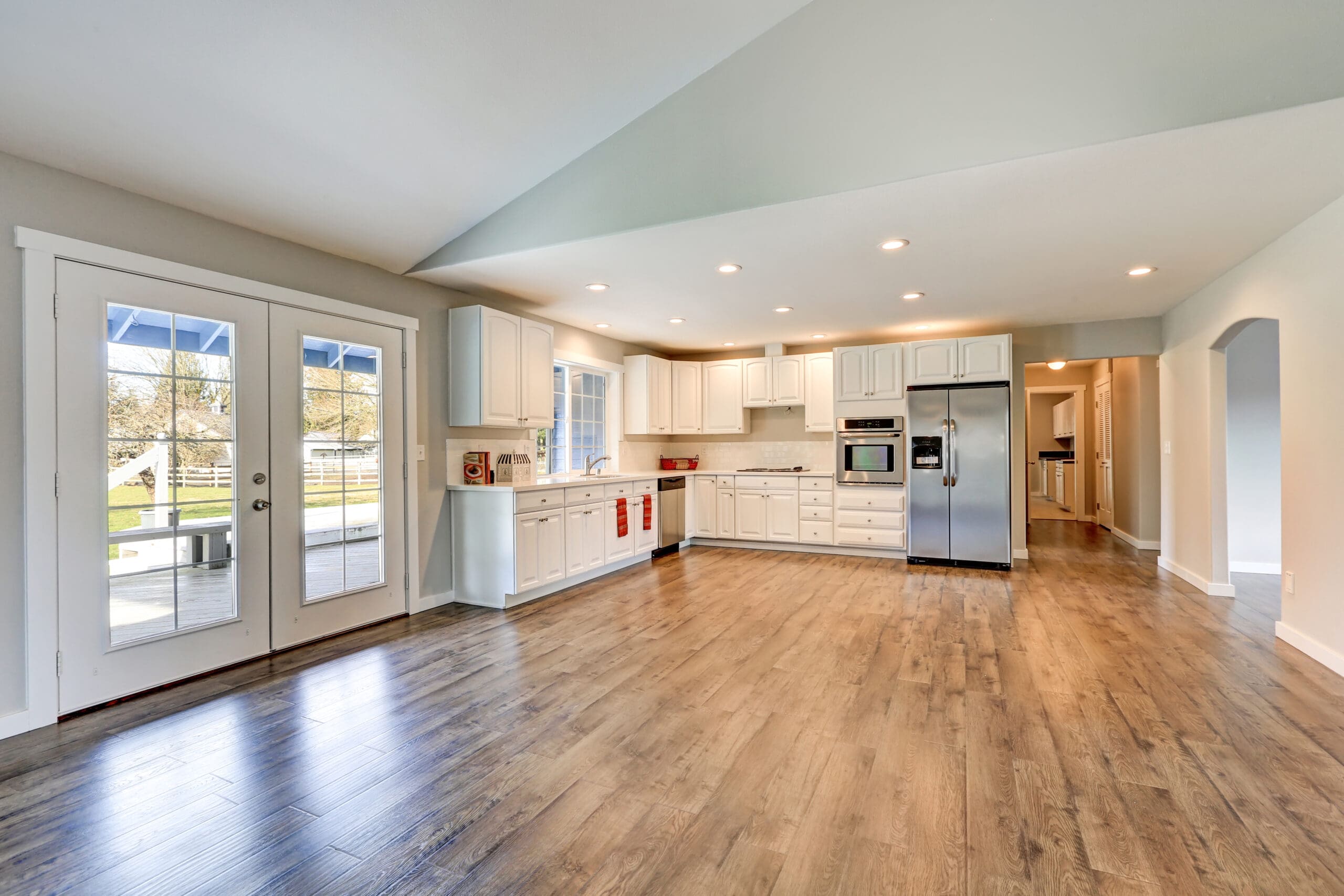 Spacious kitchen with white cabinets, stainless steel appliances, and wooden floors. Double glass doors lead to a patio. Recessed lighting brightens the room. A long hallway is visible on the right.