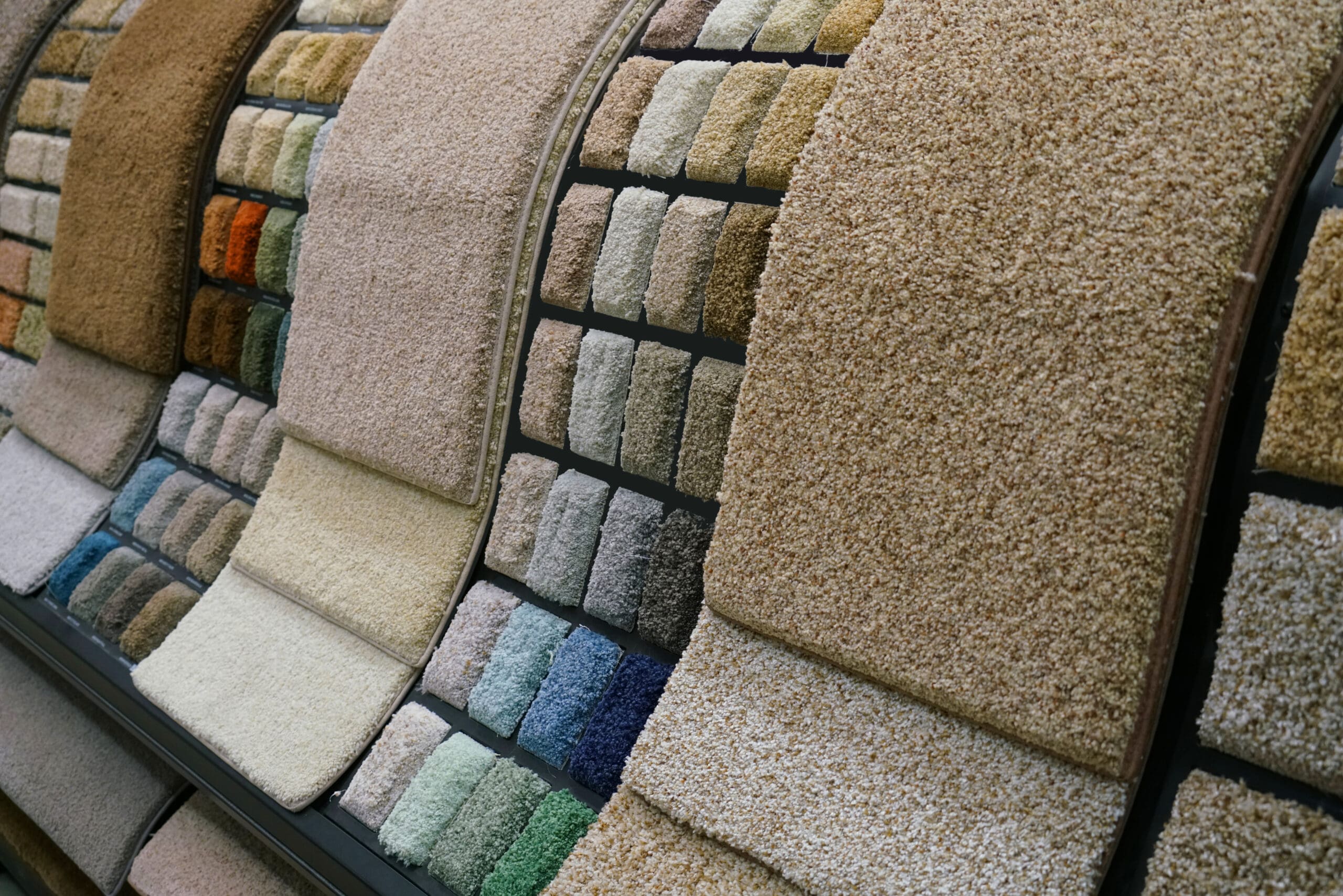 A display of various carpet samples in different colors and textures, including shades of beige, blue, green, and brown, arranged on a curved rack in a showroom.