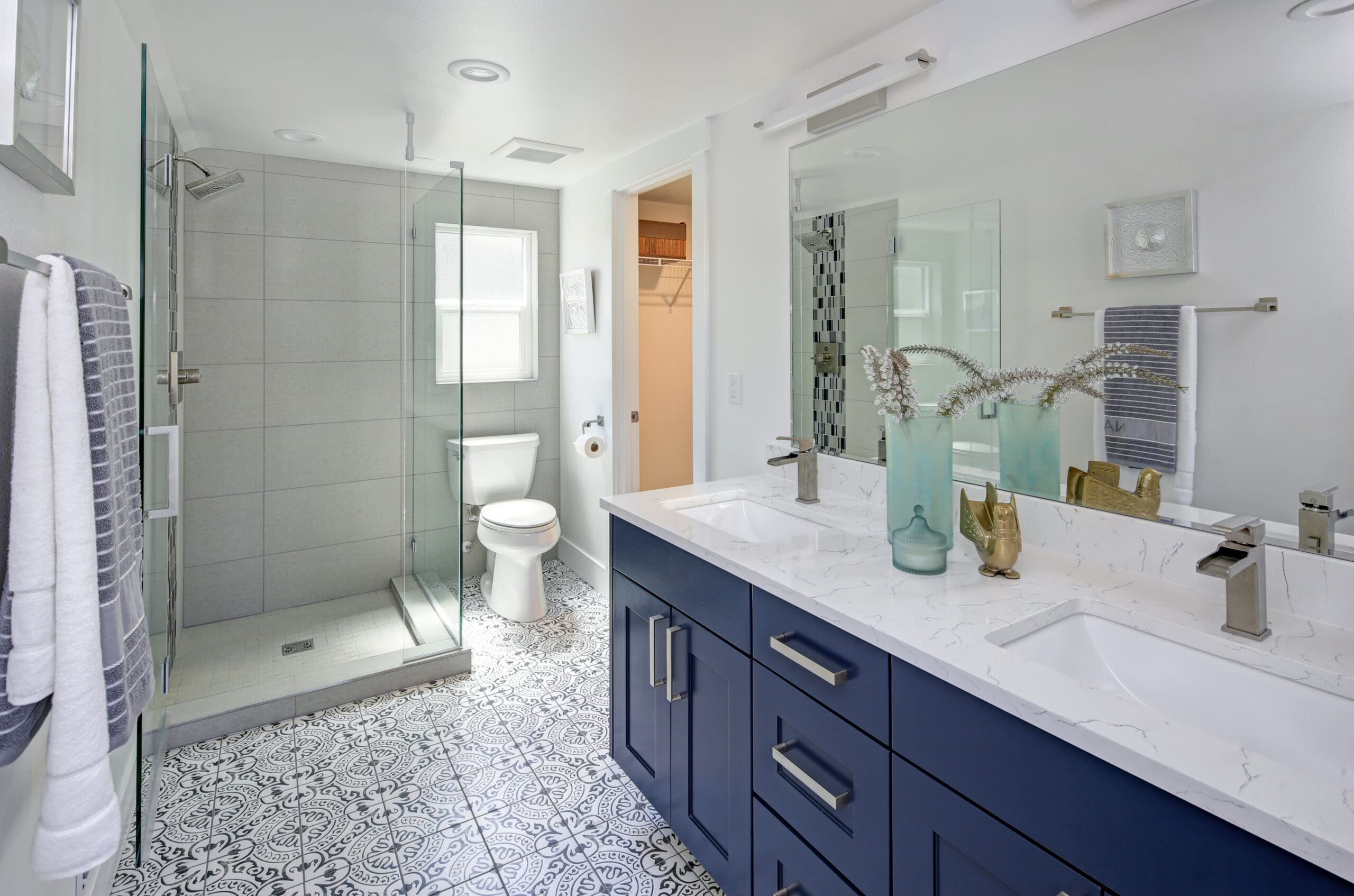 A modern bathroom featuring a glass-enclosed shower, a toilet, and a double sink vanity with blue cabinets. The floor has decorative tiles, and a large mirror adorns the wall above the sinks, complemented by contemporary decor pieces.