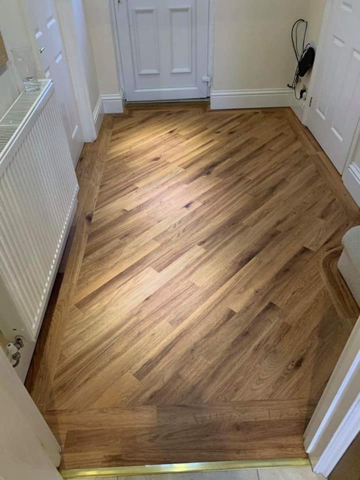 A hallway with a door at the end features a light brown wooden floor arranged in a herringbone pattern. The walls are white, and there's a white radiator on the left.