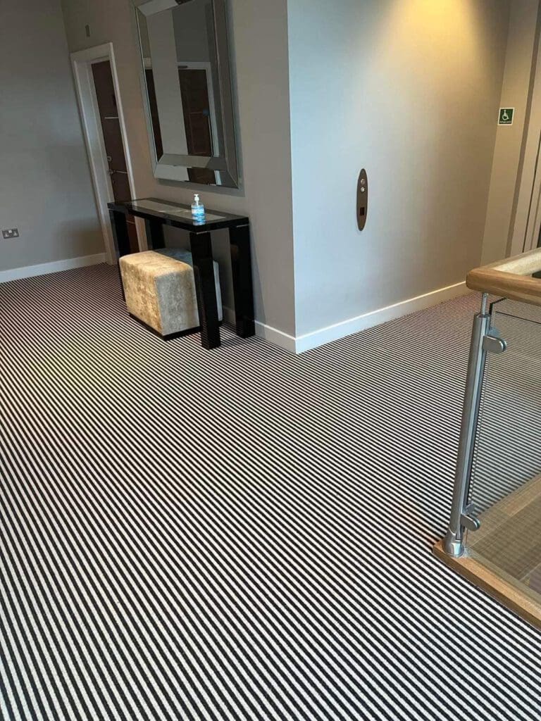 A hallway with a black and white striped carpet, a mirrored wall, and a dark wooden console table with a beige stool tucked underneath. A glass railing is visible on the right, and there's a hand sanitizer bottle on the table.