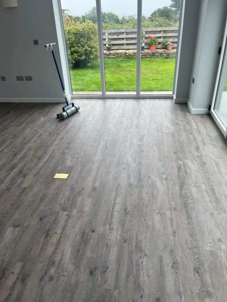 A bright room with large windows and a view of a garden. The floor is covered with wood-patterned vinyl planks. A floor roller with a handle rests to the side, and a small yellow sticky note is visible on the floor.