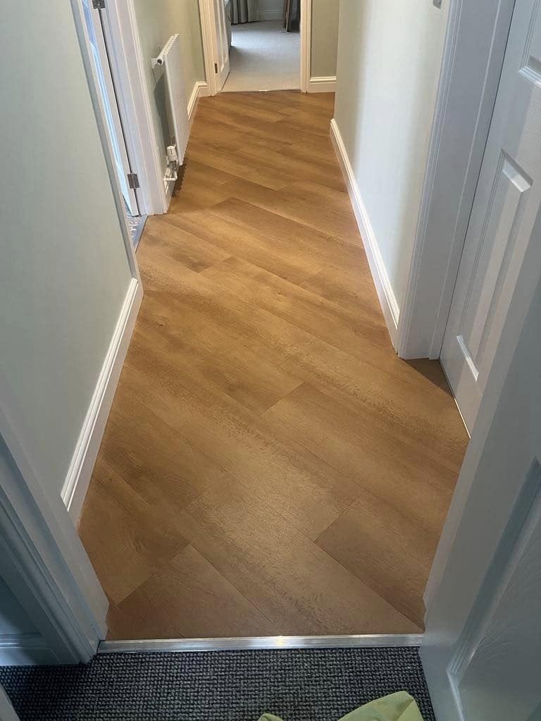 A hallway with chevron-patterned wooden flooring leads to multiple rooms with white doors. The walls are painted a light color, and a radiator is visible on the left side. The foreground shows a transition from carpet to wood.