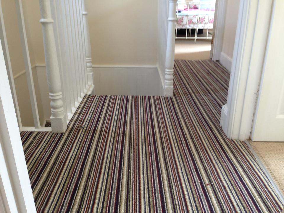 A hallway with a striped carpet in shades of brown, purple, and beige. White doors and walls line the hallway, with a staircase and wooden railings visible. Sunlight illuminates the space from a nearby room.