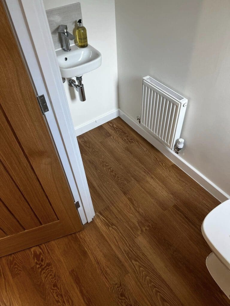 Small bathroom with wood flooring. A white sink with a soap dispenser is mounted on the wall above. A white radiator is on the opposite wall near the closed wooden door. Part of a white toilet is visible in the lower right corner.