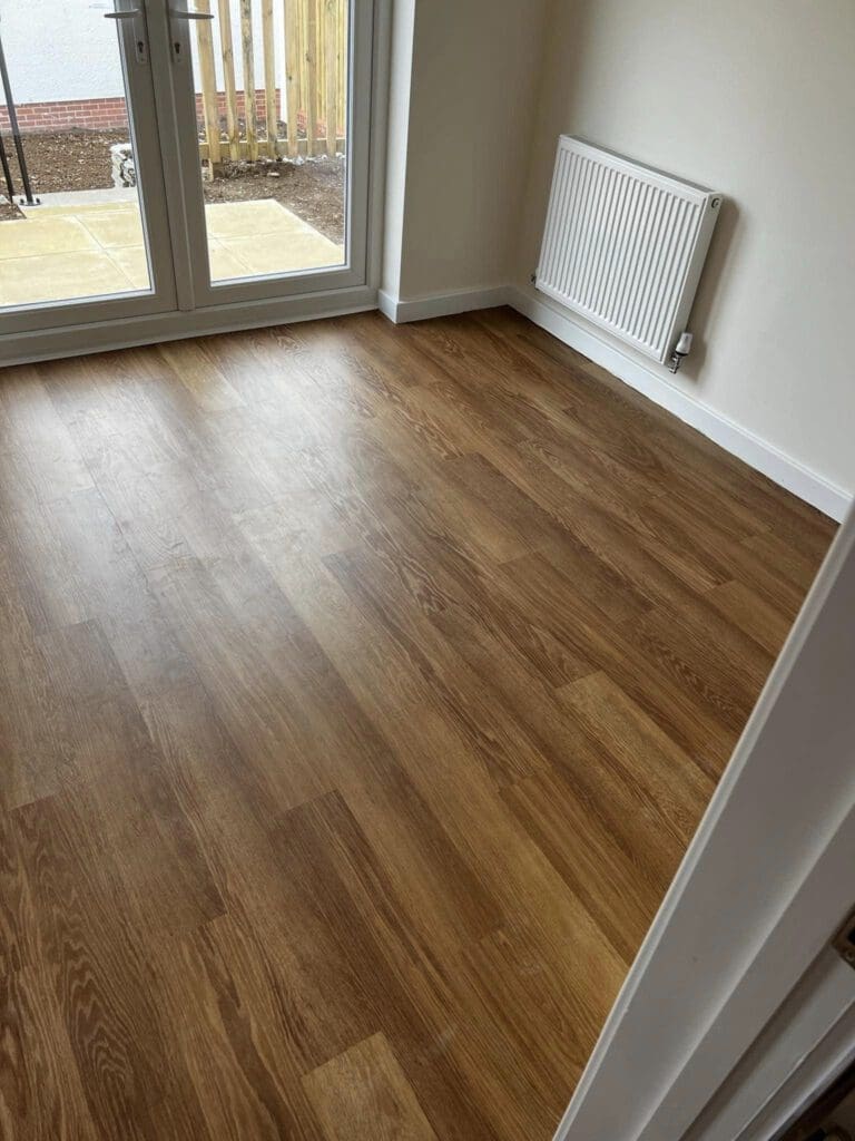 A room with wooden laminate flooring, white walls, a radiator, and a set of glass doors leading to a patio outside. The space is well-lit with natural light.