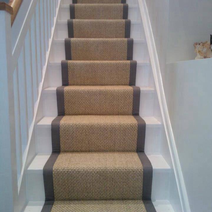 A staircase with light beige carpeting bordered by dark gray edges, running up the center of white wooden steps. The surrounding walls are also white, and a wooden handrail is visible on the left side.