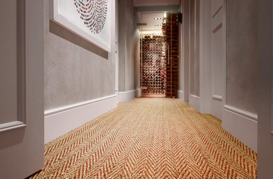 A hallway with patterned carpet leads to a well-lit area. The walls have framed artwork, and the end of the hall features a decorative panel with circular patterns in a modern design.