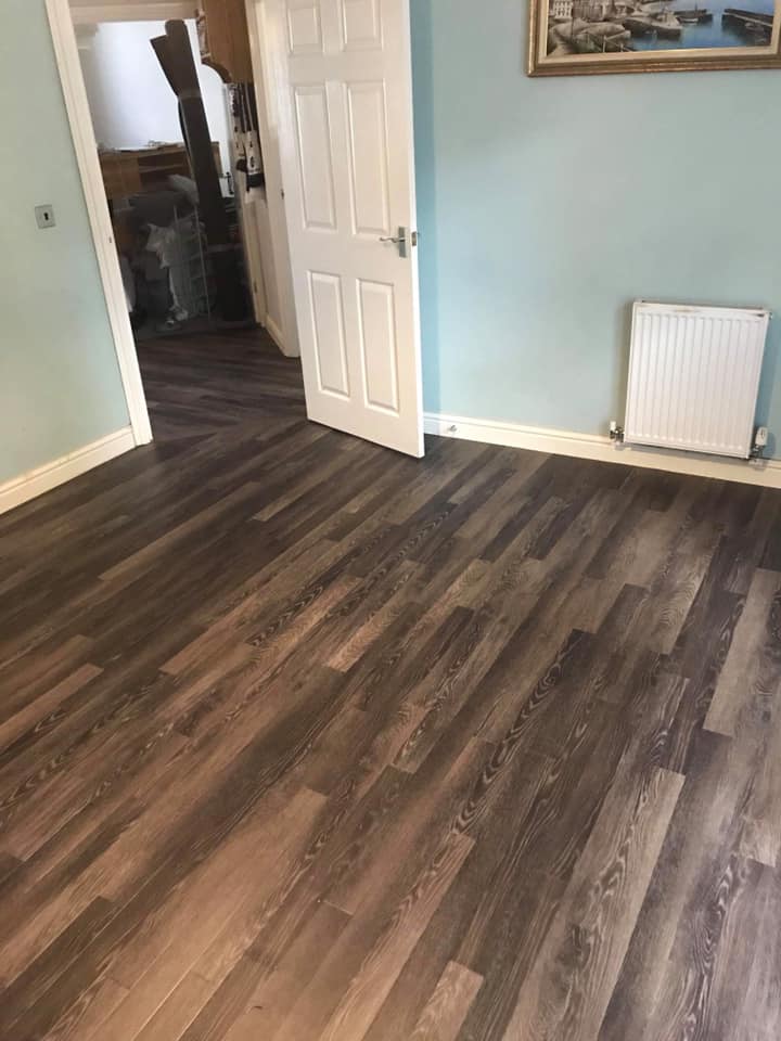 A room with light blue walls and dark wood laminate flooring. An open white door leads to another room. A white radiator is on the wall, and a framed picture hangs above it.