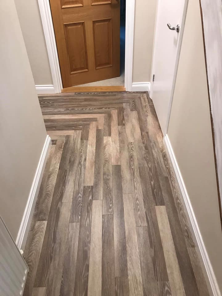 A hallway with wood-patterned vinyl flooring in a herringbone design. The walls are light-colored, and there is a wooden door slightly open at the end of the hallway with white trim, capturing a clean and modern interior look.