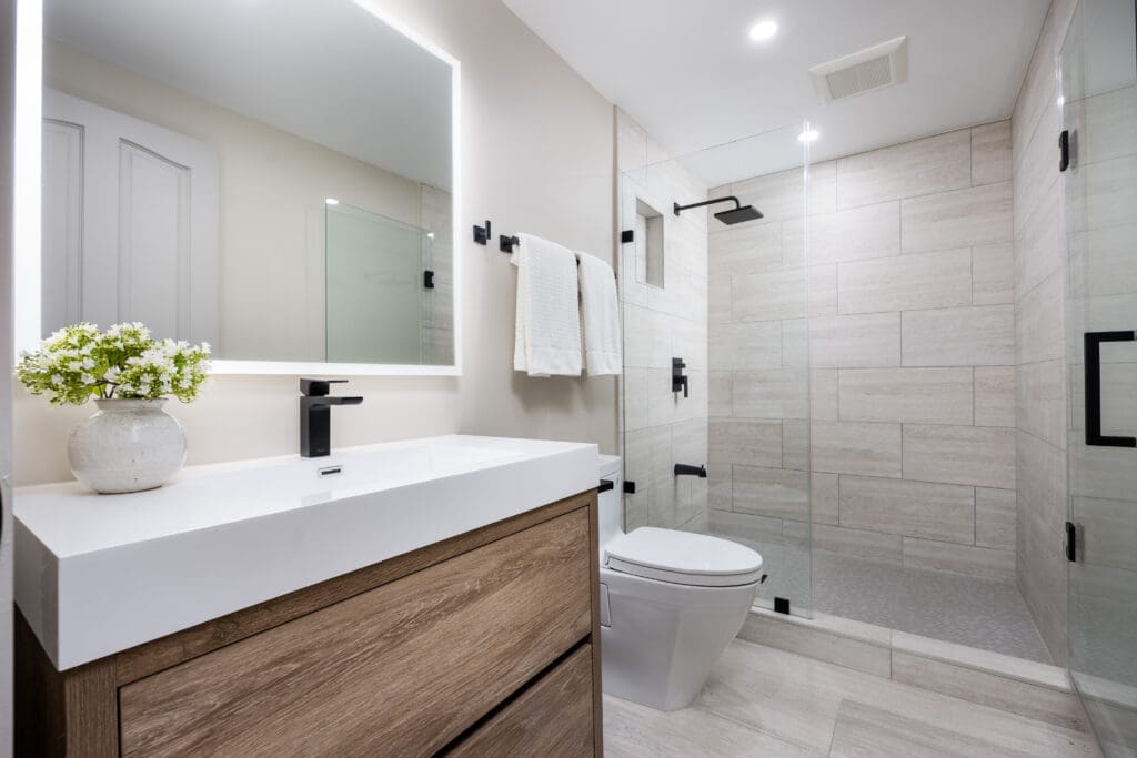 Modern bathroom with a wooden vanity and white countertop. A black faucet adorns the sink, and a potted plant sits nearby. There's a large mirror above. The shower features glass doors and beige tiles, with a black showerhead. Towels hang beside.