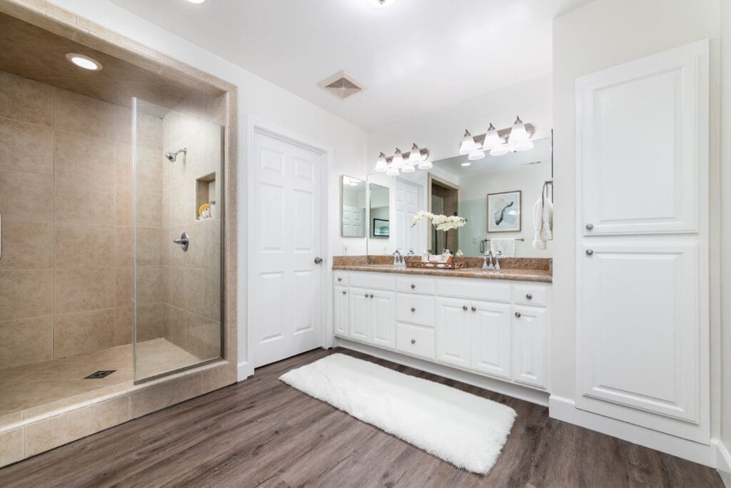 A modern bathroom with a glass-enclosed shower on the left, a large mirror above a double sink vanity with white cabinets, decorative lighting, and a fluffy white rug on wooden flooring. White walls enhance the clean, elegant look.