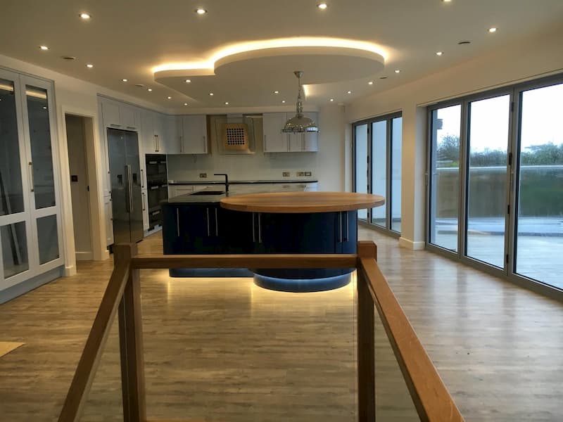 Modern kitchen with wood floors and large windows, featuring a circular island with a wooden countertop, stainless steel appliances, and recessed lighting in the ceiling. Sliding glass doors open to an outdoor view with a balcony.